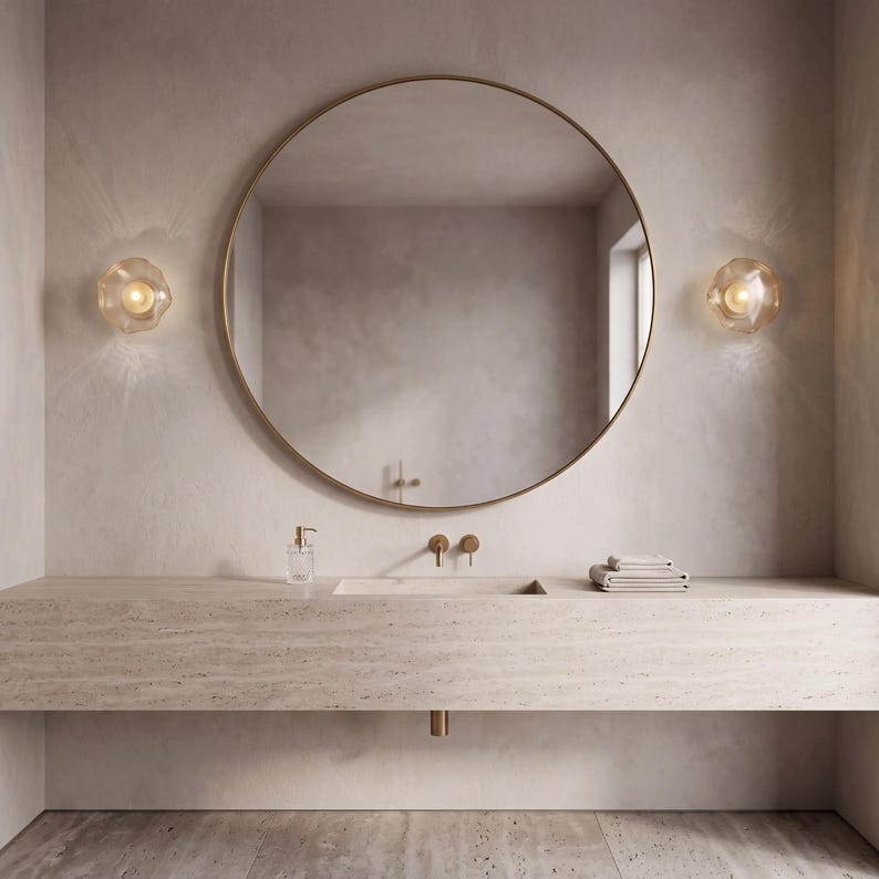 May include: A large round mirror with a thin gold frame mounted above a long, beige stone vanity. Two faceted glass wall sconces with warm glowing bulbs flank the mirror. A clear soap dispenser and a stack of folded towels sit on the counter.