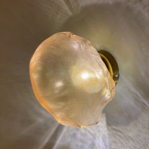 May include: A wall-mounted light fixture featuring a rounded, amber-colored mesh shade with a fine grid pattern. The shade is attached to a circular, metallic gold-toned base, and the light emits a warm glow that casts soft, textured shadows.