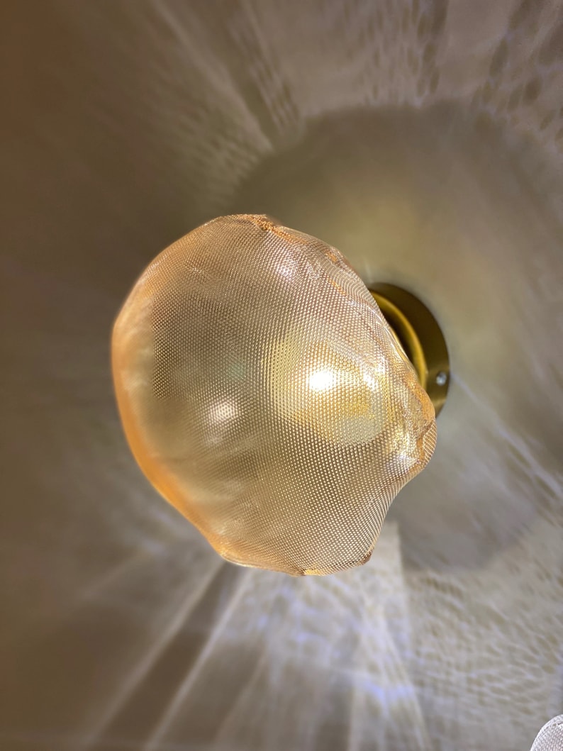 May include: A wall-mounted light fixture featuring a rounded, amber-colored mesh shade with a fine grid pattern. The shade is attached to a circular, metallic gold-toned base, and the light emits a warm glow that casts soft, textured shadows.