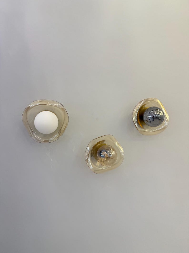 K&ouml;nnte beinhalten: Drei Wandleuchten mit unregelm&auml;&szlig;ig geformten, transparenten, bernsteinfarbenen Glasschirmen. Eine Leuchte enth&auml;lt eine mattwei&szlig;e Kugellampe, w&auml;hrend die anderen beiden kleinere, reflektierende, metallisch silberne Kugellampen enthalten.