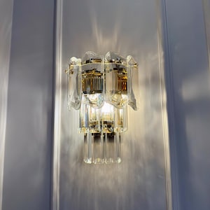 May include: A wall-mounted light fixture with a gold-toned metal frame and multiple tiers of clear, faceted crystal elements. The fixture is illuminated, casting light and shadows on the white wall. The design is ornate and decorative.