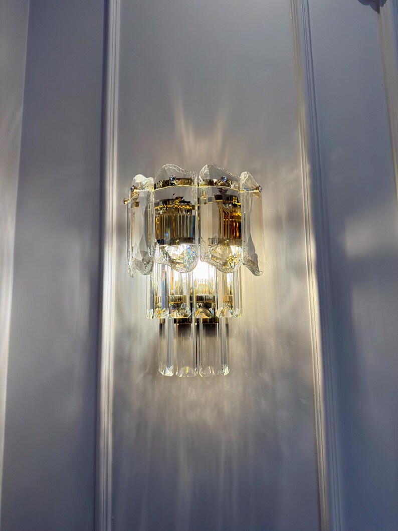 May include: A wall-mounted light fixture with a gold-toned metal frame and multiple tiers of clear, faceted crystal elements. The fixture is illuminated, casting light and shadows on the white wall. The design is ornate and decorative.