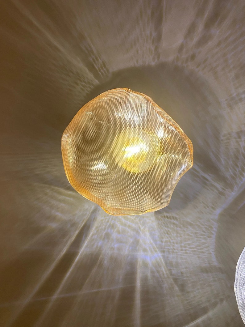 May include: A gold-toned, organic-shaped pendant light fixture with a fine mesh texture. The lamp emits a warm, glowing light that projects intricate, radiating patterns onto the surrounding area, creating a soft, diffused illumination effect.