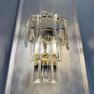 May include: A wall-mounted light fixture with a tiered design. The fixture features multiple clear, rectangular crystal elements and gold-colored metal accents. The light is turned on, illuminating the crystals and the surrounding wall.