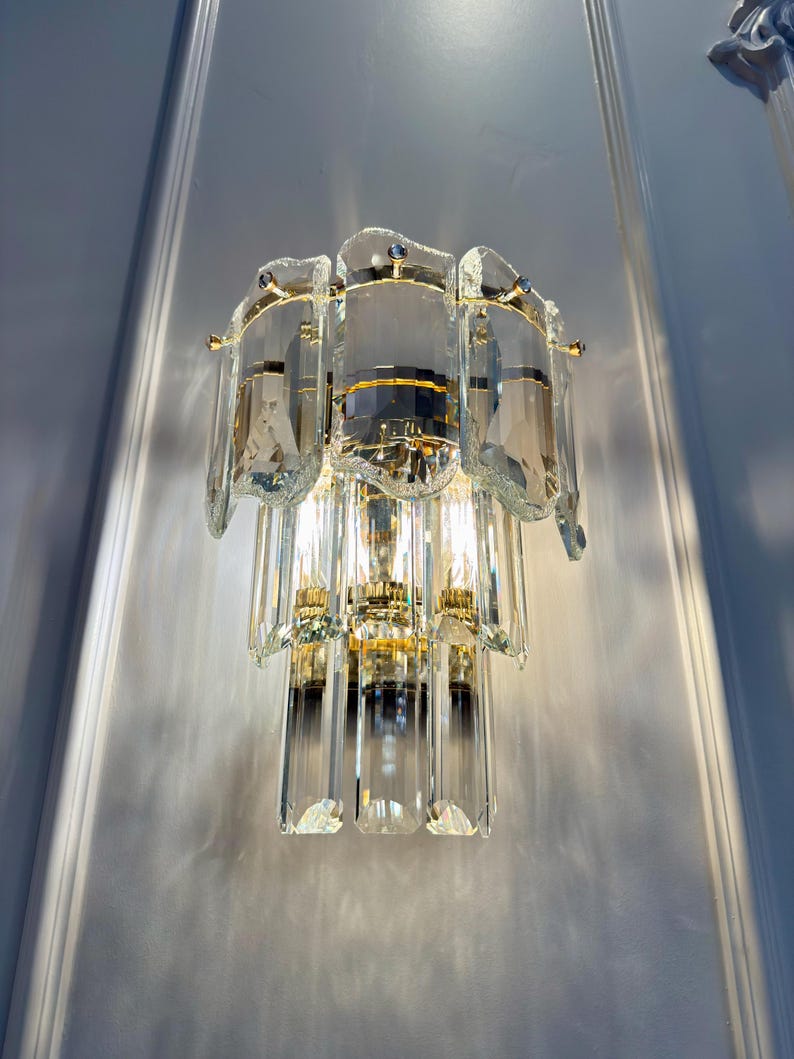 May include: A wall-mounted light fixture with a tiered design. The fixture features multiple clear, rectangular crystal elements and gold-colored metal accents. The light is turned on, illuminating the crystals and the surrounding wall.