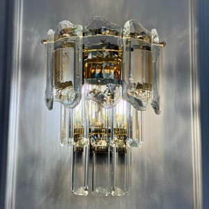 May include: A crystal wall sconce with a multi-tiered design. The sconce features clear, rectangular crystal elements and gold-toned metal accents. The light fixture is mounted on a white wall, reflecting light and creating a sparkling effect.