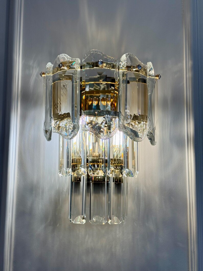 May include: A crystal wall sconce with a multi-tiered design. The sconce features clear, rectangular crystal elements and gold-toned metal accents. The light fixture is mounted on a white wall, reflecting light and creating a sparkling effect.