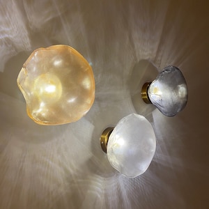 May include: Three wall-mounted light fixtures with irregular, organic-shaped glass shades. One shade is amber-colored, one is clear, and one is a smoky gray. Each lamp features a circular brass-toned base and emits a warm, glowing light.