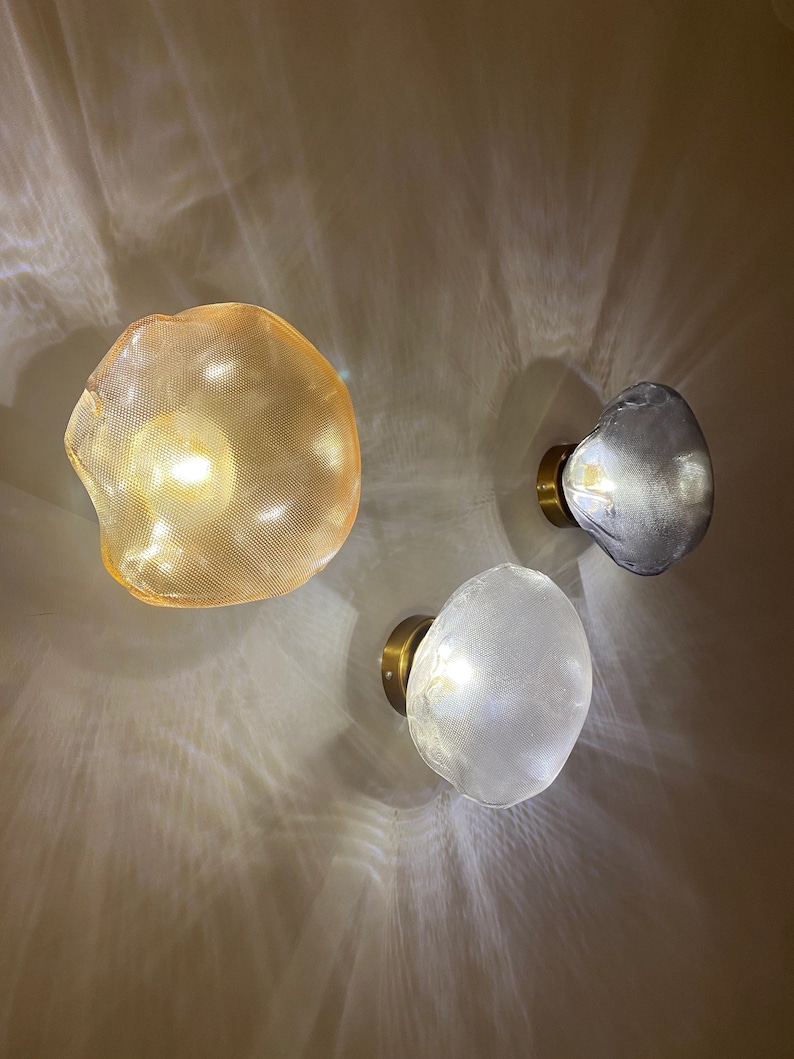 May include: Three wall-mounted light fixtures with irregular, organic-shaped glass shades. One shade is amber-colored, one is clear, and one is a smoky gray. Each lamp features a circular brass-toned base and emits a warm, glowing light.
