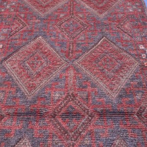 Op de afbeelding: Een rechthoekig kleed met een herhalend geometrisch patroon van grote ruiten en hoekige lijnen. Het textiel vertoont een getextureerd weefsel in diepe tinten rood, bordeaux en donkerblauw, met kleine accenten in cr&egrave;me en lichtbruin.