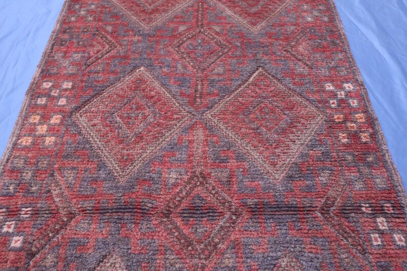 Op de afbeelding: Een rechthoekig kleed met een herhalend geometrisch patroon van grote ruiten en hoekige lijnen. Het textiel vertoont een getextureerd weefsel in diepe tinten rood, bordeaux en donkerblauw, met kleine accenten in cr&egrave;me en lichtbruin.