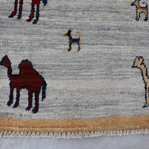May include: A light gray area rug featuring a tan border and a fringe edge. The rug displays a repeating pattern of small, stylized camels in dark brown and tan, along with small dark blue and tan animal figures scattered across the surface.