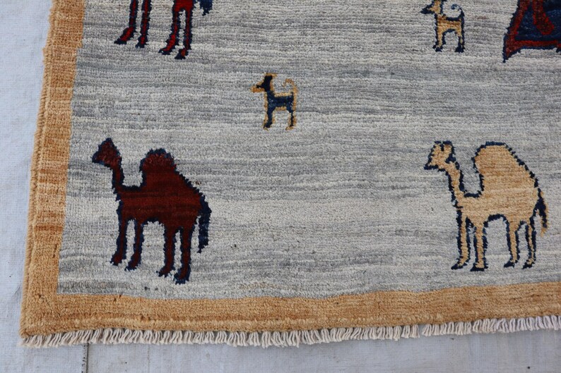 May include: A light gray area rug featuring a tan border and a fringe edge. The rug displays a repeating pattern of small, stylized camels in dark brown and tan, along with small dark blue and tan animal figures scattered across the surface.