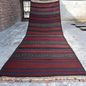 Può includere: Un lungo tappeto rettangolare con un motivo a righe nei toni del rosso, nero e blu. Il tappeto ha una trama tessuta e un bordo decorativo con frange. Il tappeto è esposto su una superficie di cemento.