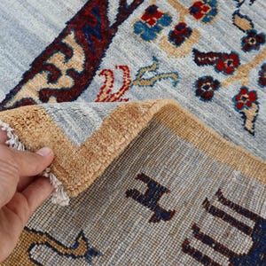 May include: A hand-knotted rug featuring a light blue field with intricate floral patterns in red, dark blue, and tan. The rug has a thick, golden-tan border and a folded corner revealing the dense, textured pile and white fringe detailing.