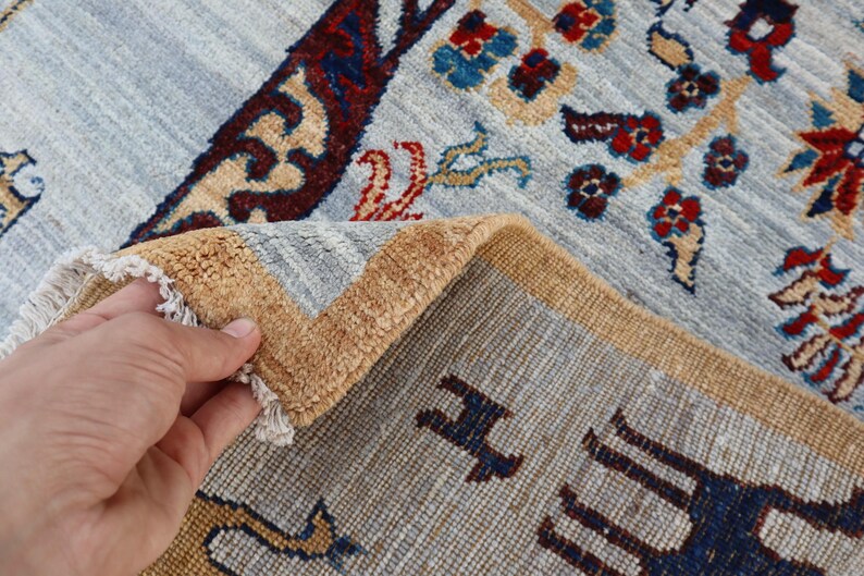 May include: A hand-knotted rug featuring a light blue field with intricate floral patterns in red, dark blue, and tan. The rug has a thick, golden-tan border and a folded corner revealing the dense, textured pile and white fringe detailing.