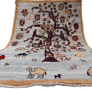 May include: A rectangular area rug with a light gray background and a tan border. The center features a large, dark-toned tree with birds perched on its branches, surrounded by various floral motifs and several camels in red, blue, and tan.