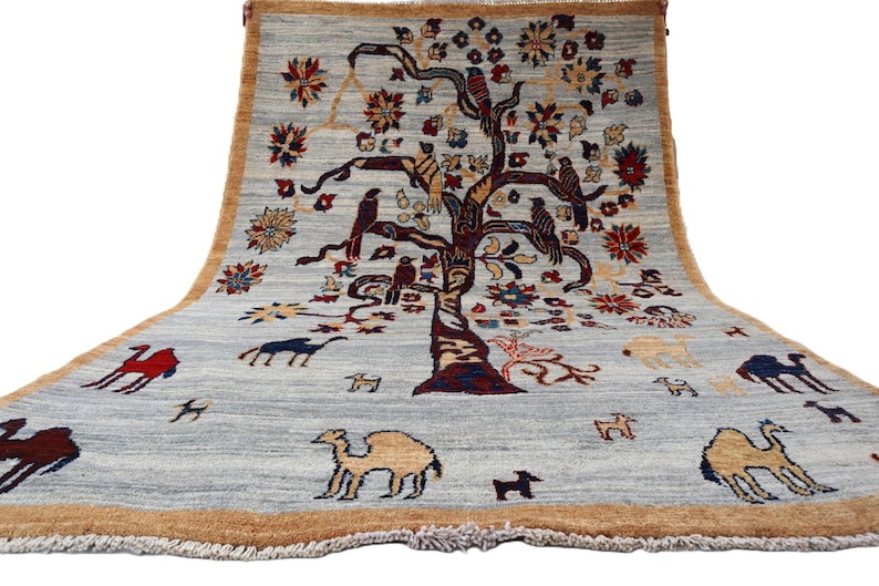 May include: A rectangular area rug with a light gray background and a tan border. The center features a large, dark-toned tree with birds perched on its branches, surrounded by various floral motifs and several camels in red, blue, and tan.