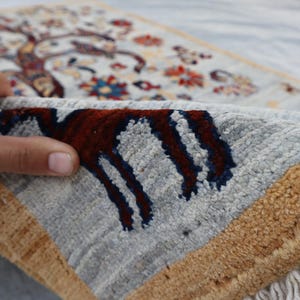 May include: A hand-knotted rug featuring a light blue field with a detailed tree of life pattern in red, blue, and gold tones. The rug has a thick, textured pile, a wide gold border, and white fringe along the edge, showcasing the dense weave.