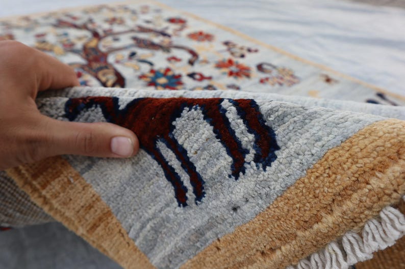 May include: A hand-knotted rug featuring a light blue field with a detailed tree of life pattern in red, blue, and gold tones. The rug has a thick, textured pile, a wide gold border, and white fringe along the edge, showcasing the dense weave.