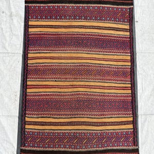 May include: A rectangular rug with a geometric pattern. The rug features alternating bands of red, orange, and dark brown. The edges have a dark brown fringe. The rug's design includes diamond and zigzag patterns.
