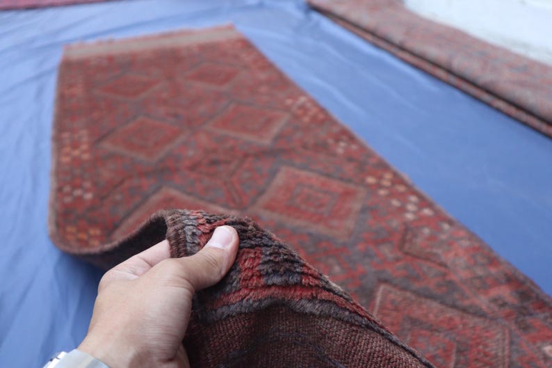 Op de afbeelding: Een lang, rechthoekig loperkleed met een dieprood en bruin kleurenpalet en een herhalend geometrisch ruitpatroon. Het kleed heeft een dikke, getextureerde pool en een zichtbare geweven rand die de constructie van het oppervlak laat zien.
