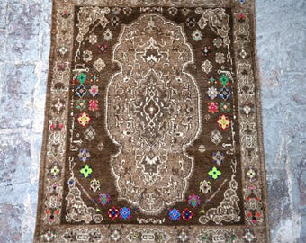Puede incluir: Una alfombra rectangular marrón con un intrincado diseño central y un borde decorativo. La alfombra presenta un patrón de medallón central en tonos más claros de marrón, rodeado por un campo marrón más oscuro. Motivos florales coloridos adornan el borde.