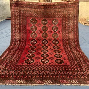 May include: A rectangular, hand-knotted rug with a red and brown geometric pattern. The rug features a central design of repeating octagons and crosses, framed by a detailed border. The rug's fringe is visible along the bottom edge.