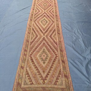 May include: A long, narrow runner rug with a repeating diamond pattern in shades of pink, beige, and olive green. The rug has a fringed edge and a geometric design. The rug is likely made of woven fabric.