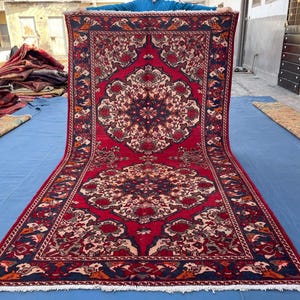 May include: A large, rectangular red rug with an intricate floral pattern in cream, navy, and brown. The rug features a central medallion design and a detailed border. The rug is displayed outdoors on a blue surface.