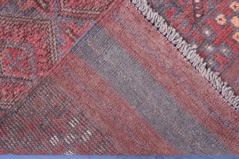 Op de afbeelding: Een geweven tapijt met een geometrisch patroon in dieprood en leiblauw, met een omgevouwen hoek die de onderkant laat zien. Het textiel vertoont een dichte pool, ingewikkelde ruitmotieven en een gefranjerde rand met zichtbare garenstructuur.