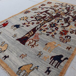 May include: A rectangular area rug with a light gray background and a tan border. The rug features a large central tree with dark brown branches, colorful floral motifs, and various animal figures including camels in red, blue, and tan.