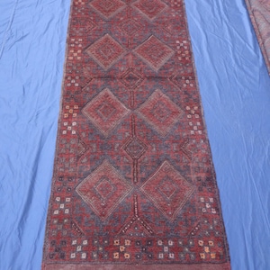 Op de afbeelding: Een rechthoekig loperkleed in een dieprode en donkerblauwe kleurstelling. Het ontwerp bevat een reeks grote, herhalende ruitvormen over de lengte, geaccentueerd door kleine geometrische motieven en franjes aan beide uiteinden.