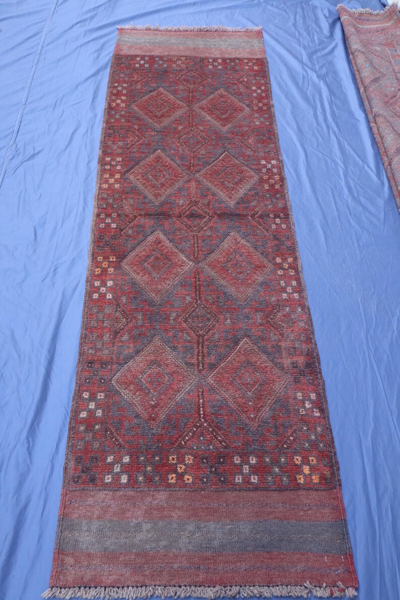 Op de afbeelding: Een rechthoekig loperkleed in een dieprode en donkerblauwe kleurstelling. Het ontwerp bevat een reeks grote, herhalende ruitvormen over de lengte, geaccentueerd door kleine geometrische motieven en franjes aan beide uiteinden.
