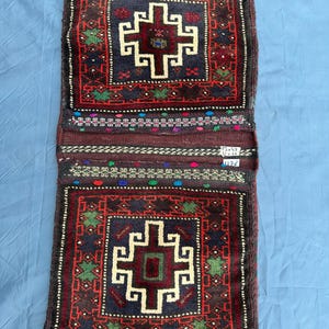 Pode incluir: Um têxtil retangular tecido com um padrão geométrico repetitivo em vermelho, azul marinho e creme. O design apresenta uma forma central em forma de cruz e é emoldurado por uma borda de formas e pontos menores. O têxtil é provavelmente um tapete ou uma peça decorativa.