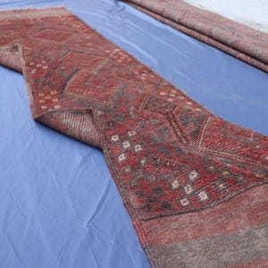 Op de afbeelding: Een lang, smal loperkleed met een kleurenpalet van dieprood en donker antraciet. Het textiel vertoont een herhalend geometrisch patroon van ruiten en kleine vierkante motieven, met een getextureerd, geweven oppervlak en franjes aan de uiteinden.