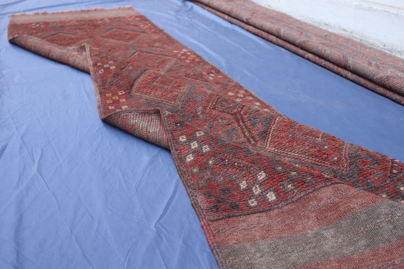 Op de afbeelding: Een lang, smal loperkleed met een kleurenpalet van dieprood en donker antraciet. Het textiel vertoont een herhalend geometrisch patroon van ruiten en kleine vierkante motieven, met een getextureerd, geweven oppervlak en franjes aan de uiteinden.