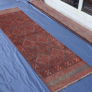 Op de afbeelding: Een lang, rechthoekig loperkleed in dieprode en bruine kleurtinten. Het centrale veld is versierd met een herhalend patroon van grote ruiten en kleinere geometrische motieven, afgewerkt met franjes aan de uiteinden en brede randen.