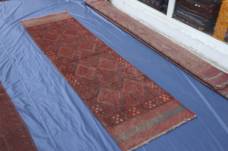 Op de afbeelding: Een lang, rechthoekig loperkleed in dieprode en bruine kleurtinten. Het centrale veld is versierd met een herhalend patroon van grote ruiten en kleinere geometrische motieven, afgewerkt met franjes aan de uiteinden en brede randen.