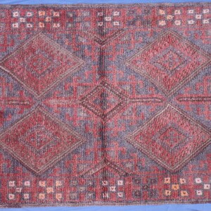 Op de afbeelding: Een rechthoekig vloerkleed met een herhalend geometrisch patroon van grote ruiten in tinten dieprood en gedempt paars. Het ontwerp bevat ingewikkelde lineaire details en kleine vierkante accenten in lichtere tinten over het getextureerde oppervlak.