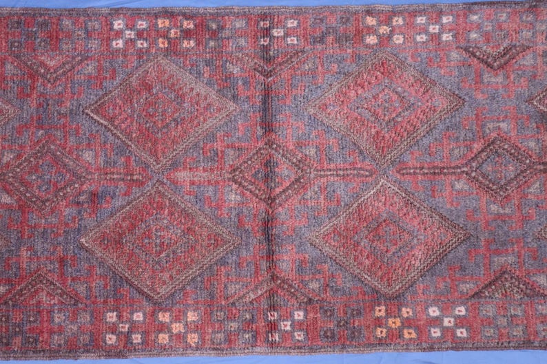 Op de afbeelding: Een rechthoekig vloerkleed met een herhalend geometrisch patroon van grote ruiten in tinten dieprood en gedempt paars. Het ontwerp bevat ingewikkelde lineaire details en kleine vierkante accenten in lichtere tinten over het getextureerde oppervlak.
