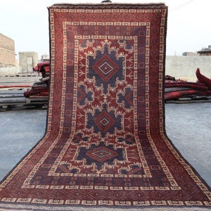 Pode incluir: Um tapete retangular grande com um padrão geométrico em tons de bordô, azul marinho e bege. O tapete apresenta um motivo central de diamante e desenhos de borda intrincados. O tapete é exibido ao ar livre, mostrando seu tamanho e detalhes.