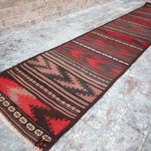 Puede incluir: Una alfombra larga y rectangular con un patrón geométrico. La alfombra presenta una paleta de colores rojo, marrón y negro, con un borde con flecos. El diseño incluye rayas y formas estilizadas.