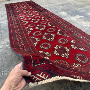 Pode incluir: Um tapete vermelho retangular e longo com um padrão geométrico. O tapete apresenta um design repetitivo de octógonos e quadrados em creme e castanho. As bordas têm uma borda azul escura e franjas creme. O tapete é exibido em uma superfície de concreto.