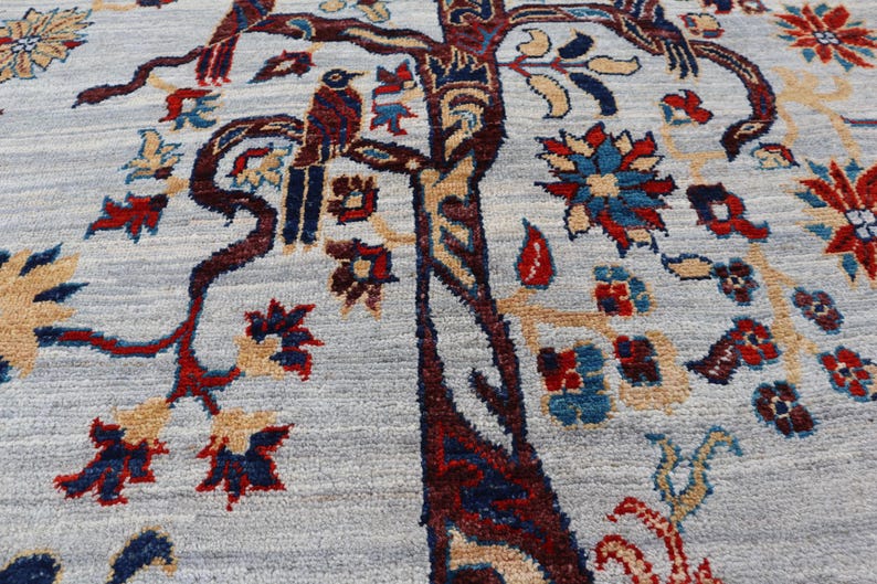 May include: A light blue area rug featuring a detailed woven pattern of a large central tree with dark red and brown bark. The design includes stylized birds perched on branches, surrounded by intricate floral motifs in red, blue, and tan.