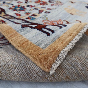 May include: A hand-knotted wool rug featuring a light gray background with a central tree motif in dark red, blue, and orange. The rug has a thick tan border and a folded corner revealing the dense pile, intricate animal figures, and white fringe.