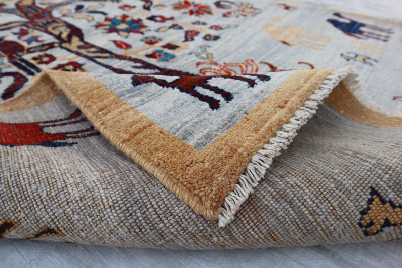 May include: A hand-knotted wool rug featuring a light gray background with a central tree motif in dark red, blue, and orange. The rug has a thick tan border and a folded corner revealing the dense pile, intricate animal figures, and white fringe.