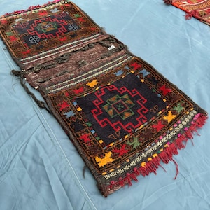 Pode incluir: Bolsa têxtil tecida à mão com um padrão geométrico em vermelho, amarelo, azul e verde sobre um fundo castanho escuro. A bolsa tem formato retangular com aba e franjas. O tecido é provavelmente de lã.