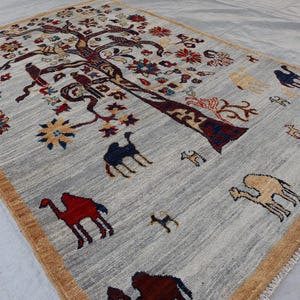 May include: A rectangular area rug with a light gray field and a tan border. The rug features a large central tree motif with dark brown branches, colorful floral blossoms, and various animal figures including camels in red, blue, and tan.