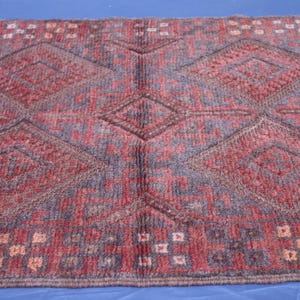 Op de afbeelding: Een rechthoekig vloerkleed met een herhalend patroon van grote ruiten in tinten dieprood en gedempt blauw. Het ontwerp bevat kleinere geometrische motieven en een rand met een raster van rode, blauwe en cr&egrave;mekleurige vierkantjes.